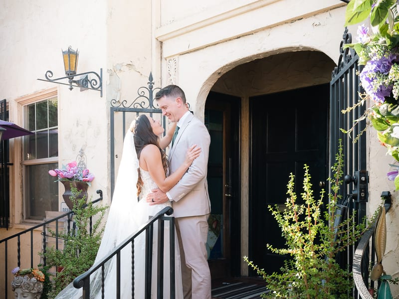 Man and woman in bridal attire standing on a stoop leading up to rustic building.