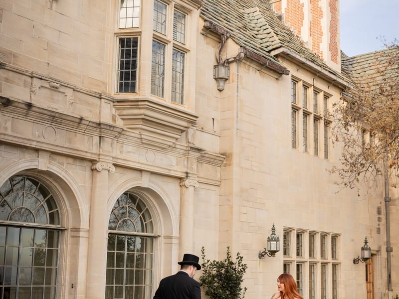 A man in a black suit with tails, a top hat, and a cane dancing with a woman with long red hair and long white bridal gown and tattoos. They are standing in a court yard at Greystone Mansion in Los Angeles, CA.