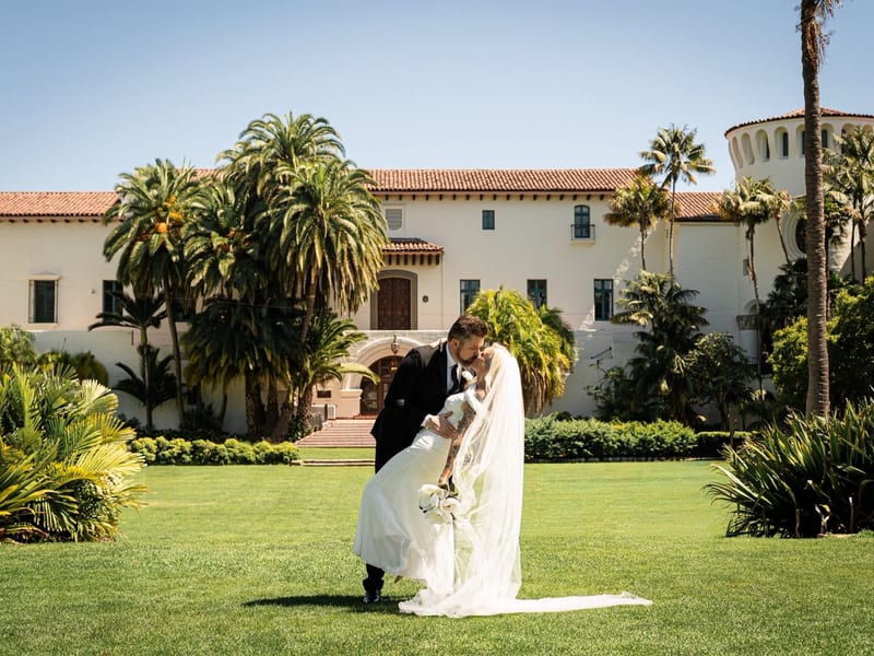 Man and woman in bridal attire. He is dipping and kissing her. They are standing on a green lawn with the Santa Barbara Court House in the distance. It's a Spanish style building.