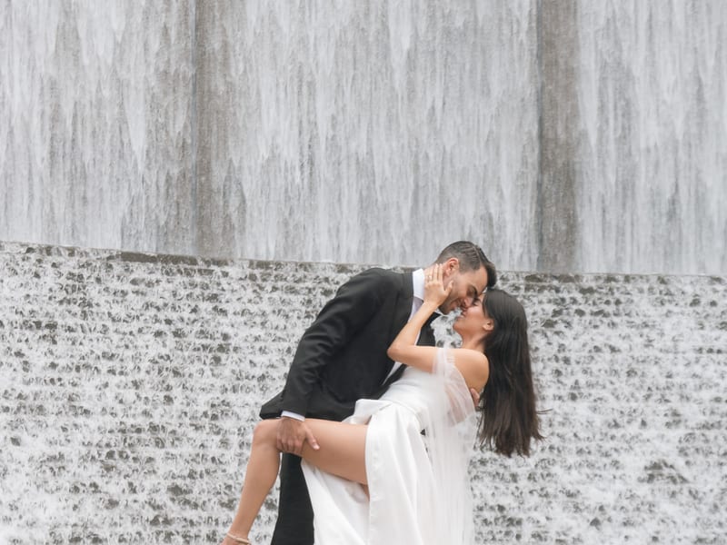 A man in black tuxedo dips a woman in a long white bridal gown in front of the Water Wall in Houston. Water is flowing down the tall wall like a waterfall.